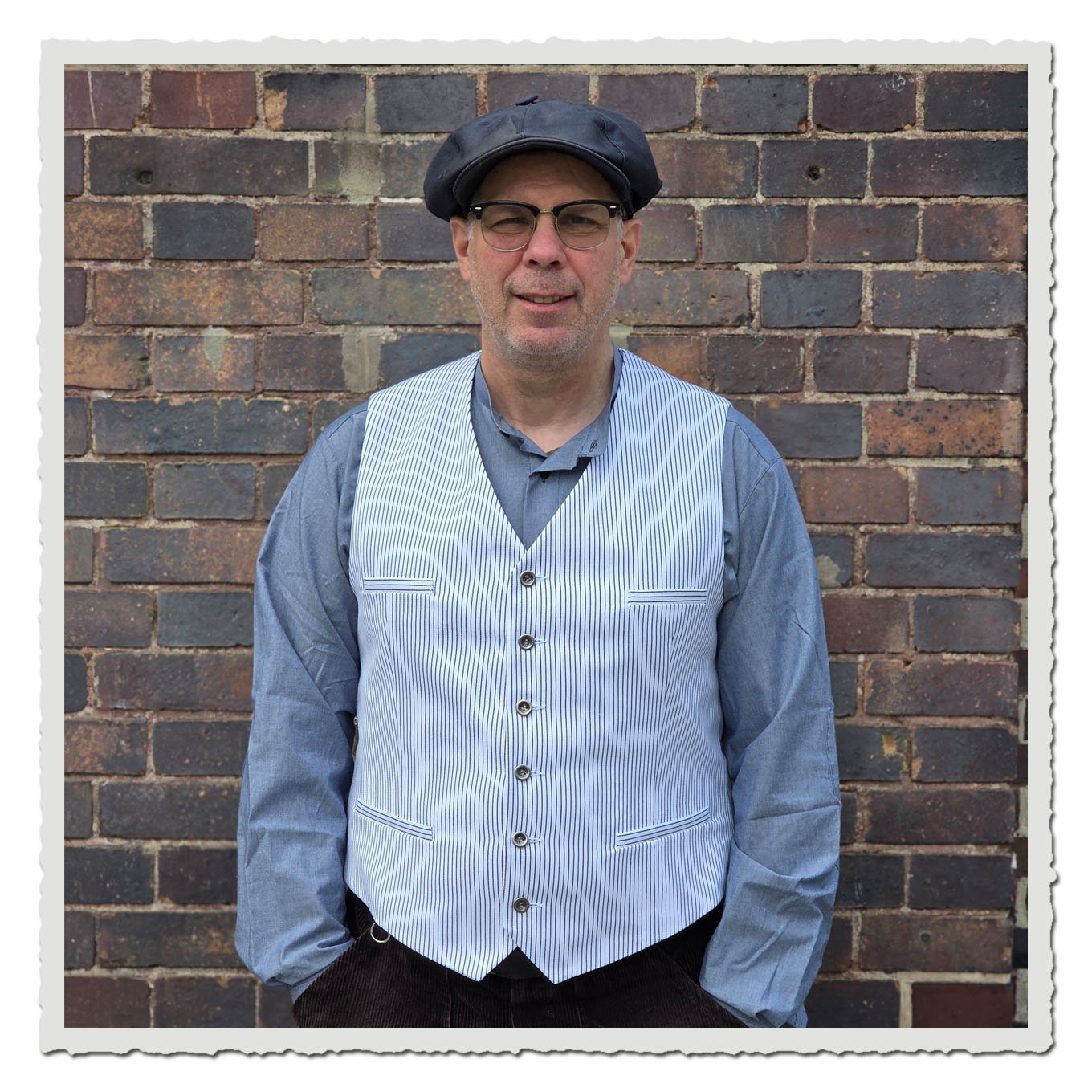 Mann mit heller gestreifter Weste und dunkler Mütze, Frontalansicht vor einer braunen Backsteinwand.